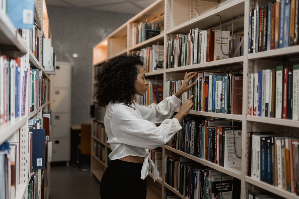 A woman stands touching books on a library shelf as if trying to find a particular one.