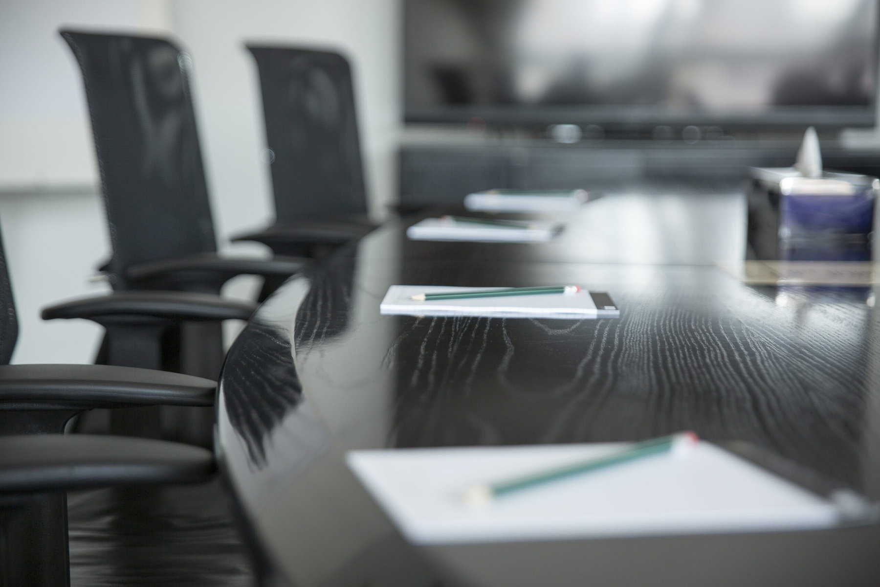 Clip boards with paper and pencils are sitting in front of chairs on a dark conference table.