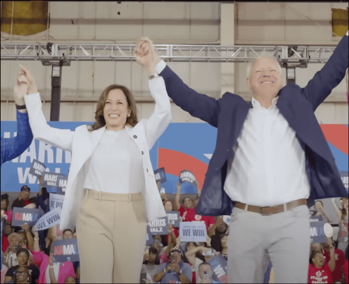 VP Kamala Harris and Tim Walz raise clasped hands in front of a crowd.