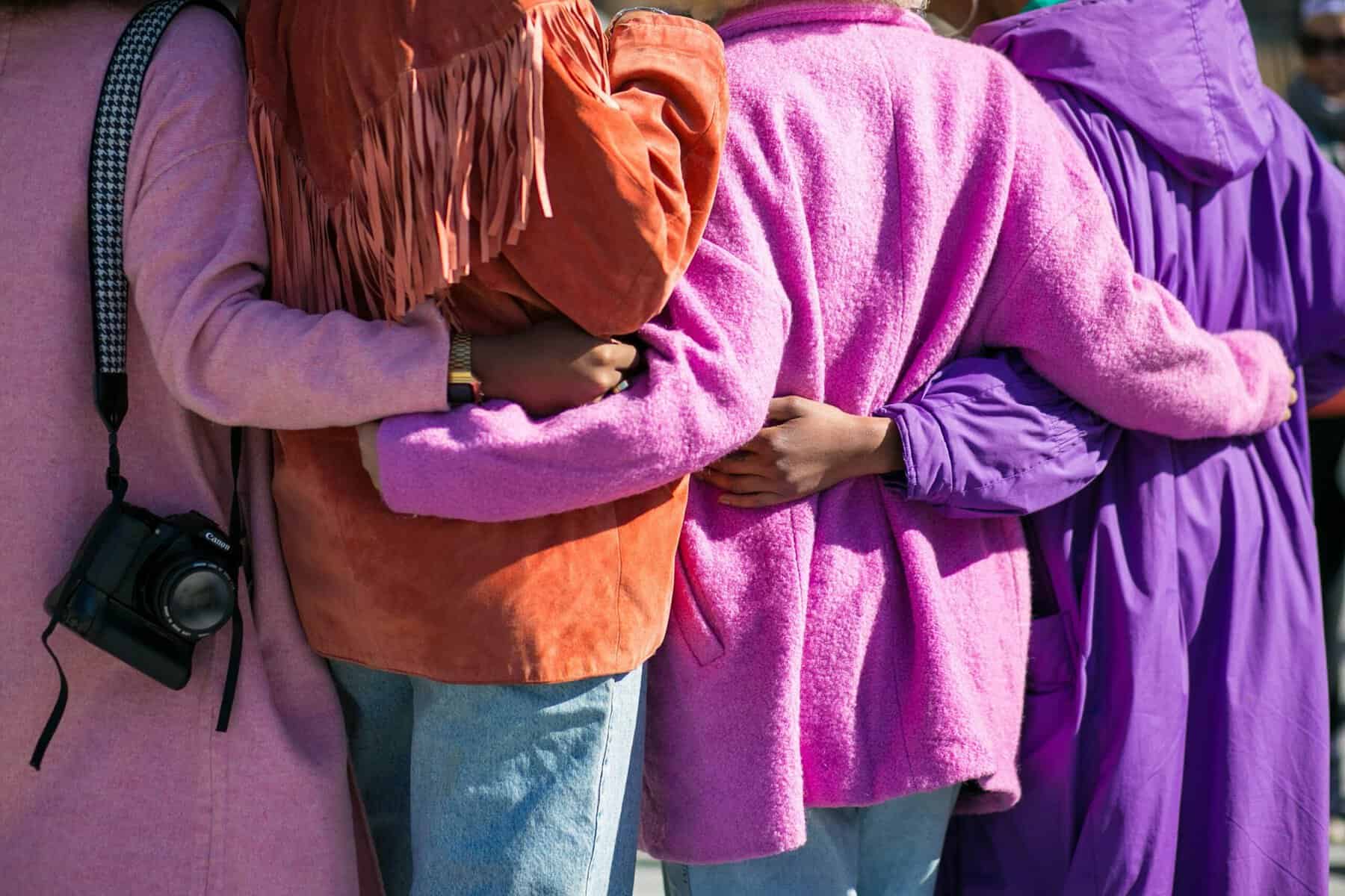 Black women are shown from the back wearing pink and orange with arms around each other's waist.