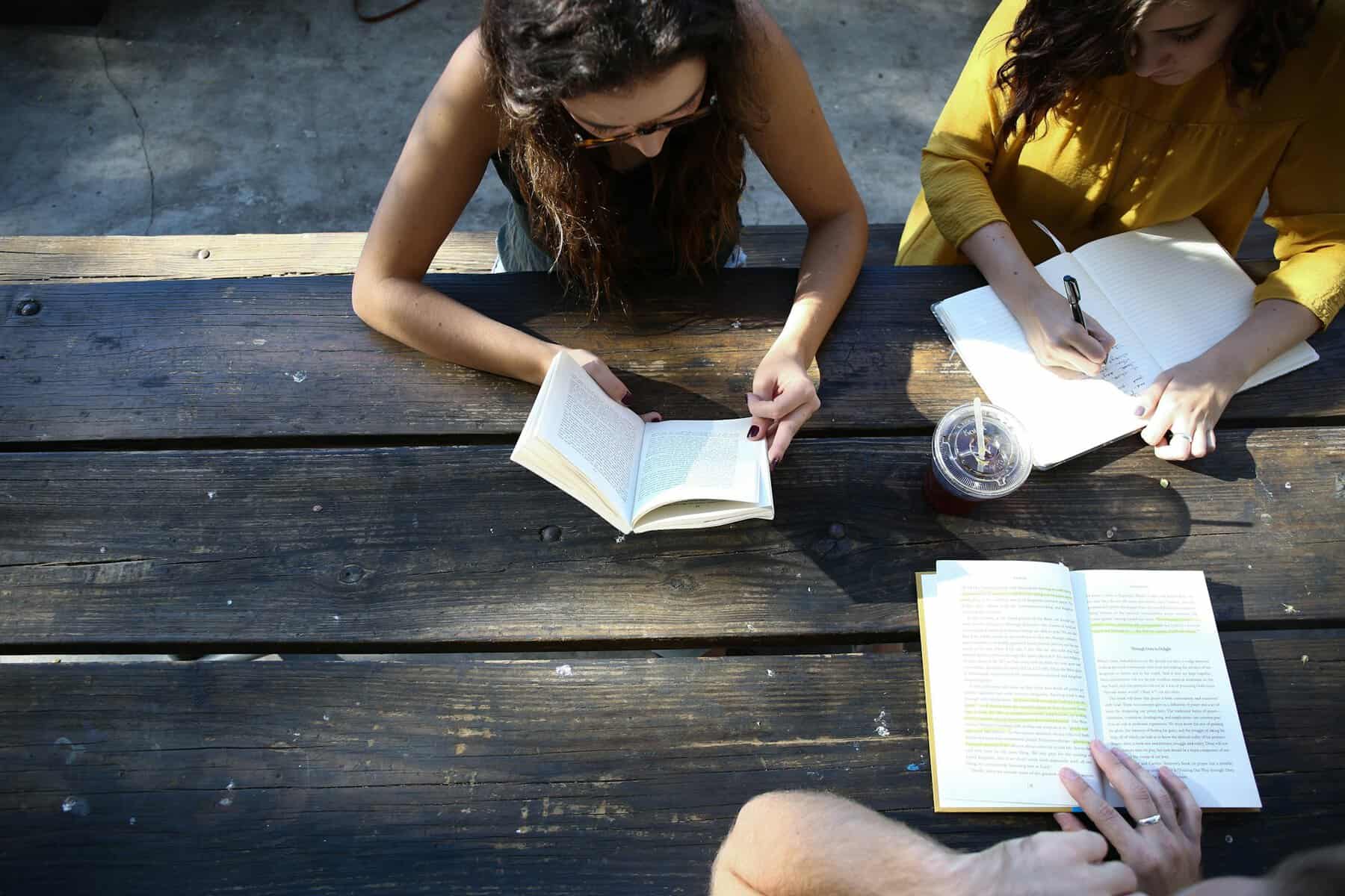 A group of people sitting at a picnic table reading books.