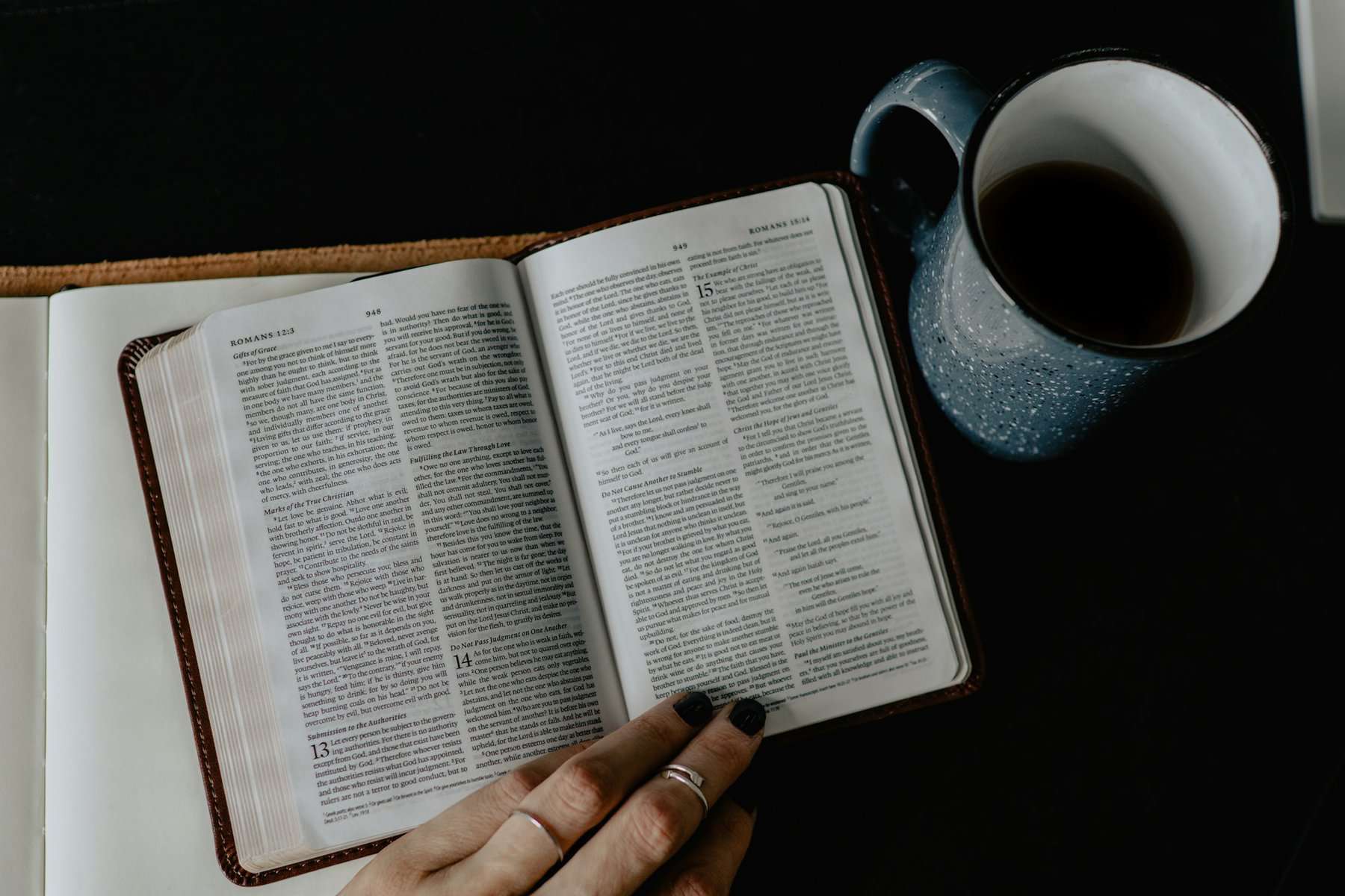 A Bible sits open to Romans with a cup of coffee and a woman's hands visible.