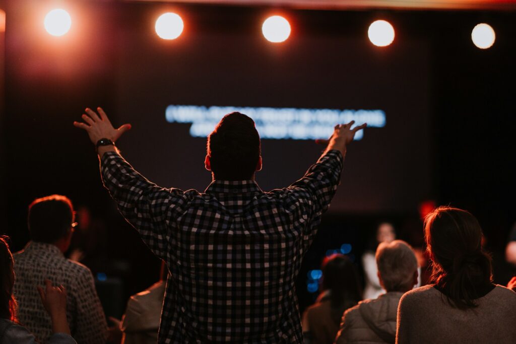 A man in a plaid shirt seen from the back lifting his hands as he sings.