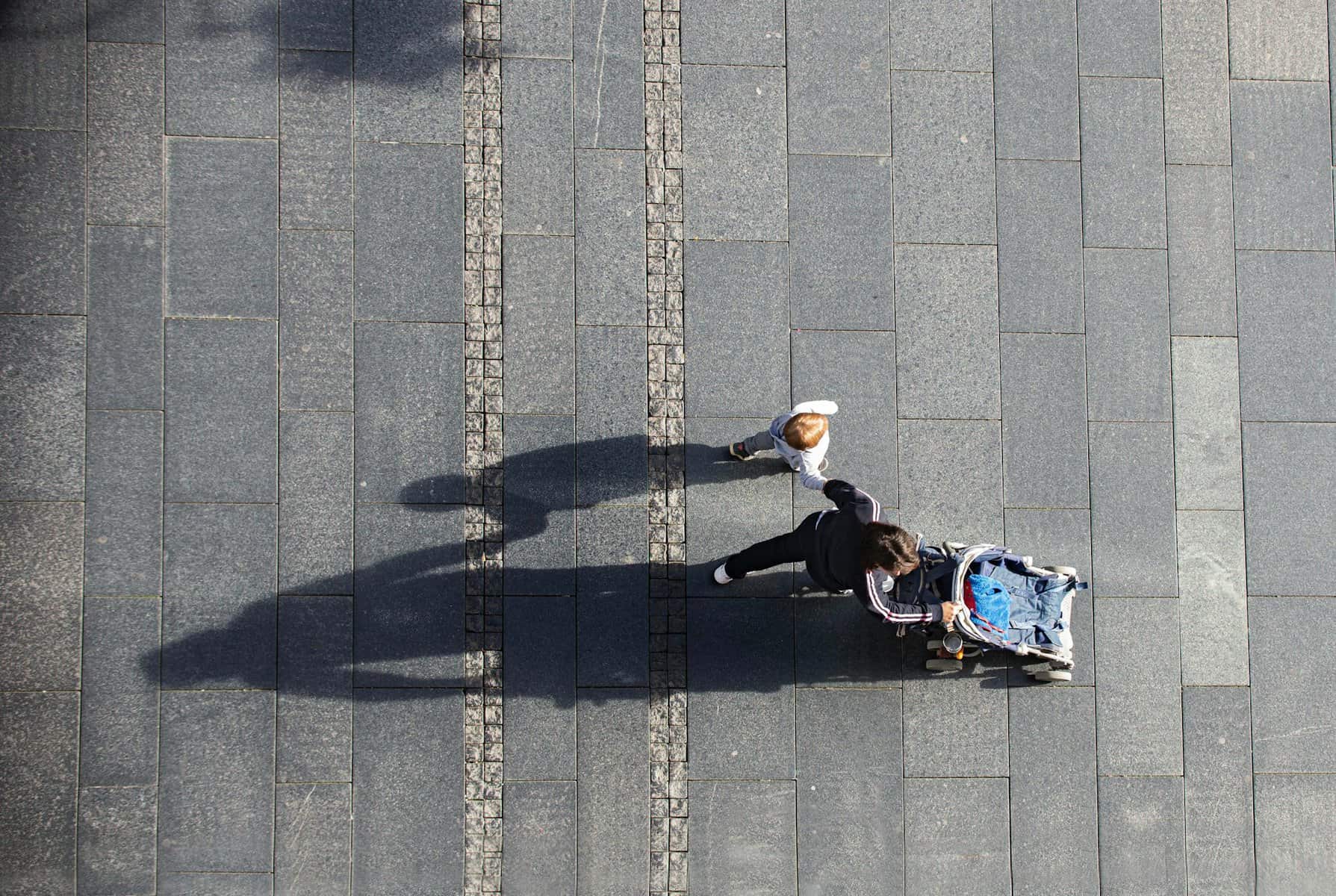 A woman is seen from high above pushing a stroller and holding a toddler's hand.