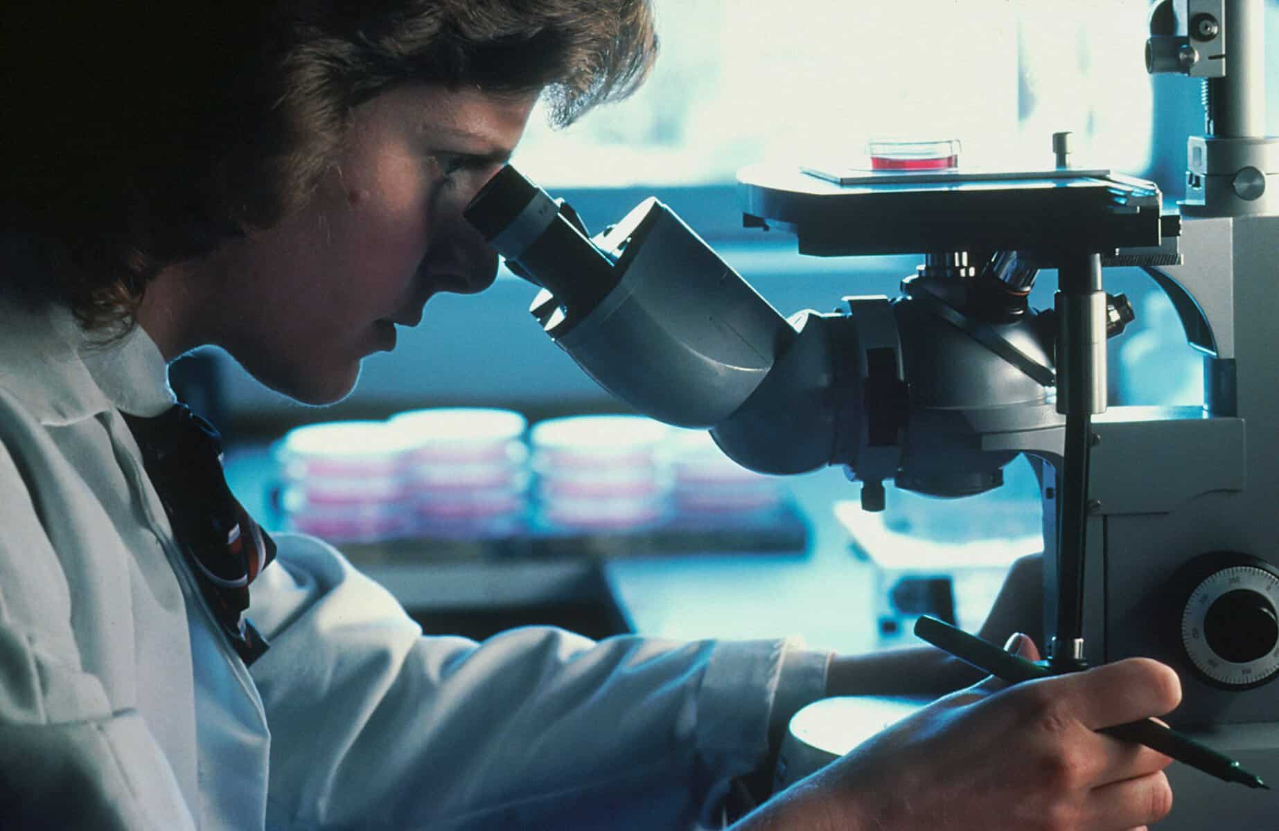 A woman in a white lab coat stares intently into a microscope.