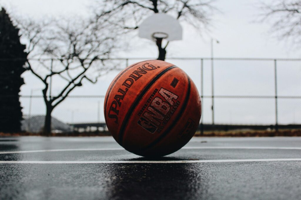 A worn basketball sits on a wet outside court with the hoop behind it.