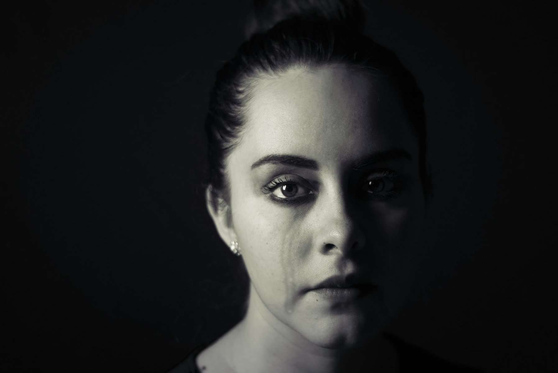 A woman in this black and white image is broad lit and stares ahead blankly with a tear running down her cheek.