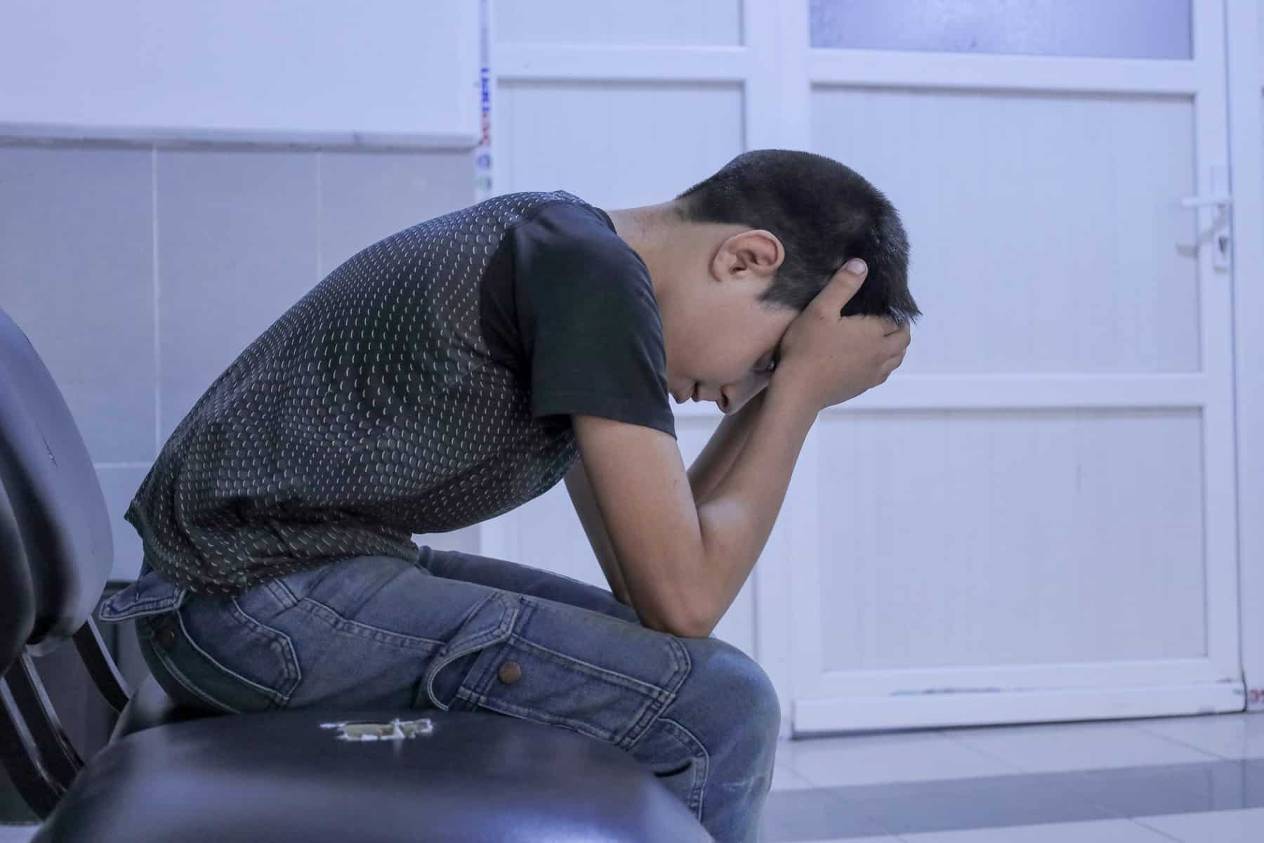 A boy sits in a gloomy room with his head in his hands.