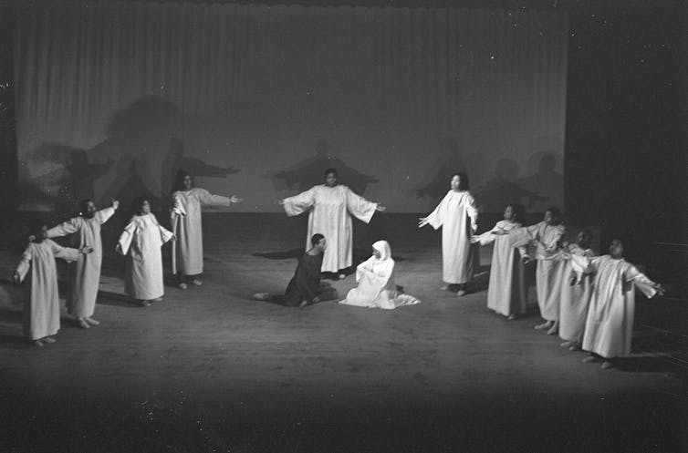 A dozen performers in white robes stand with arms outstretched around a couple sitting on the floor of an empty stage.