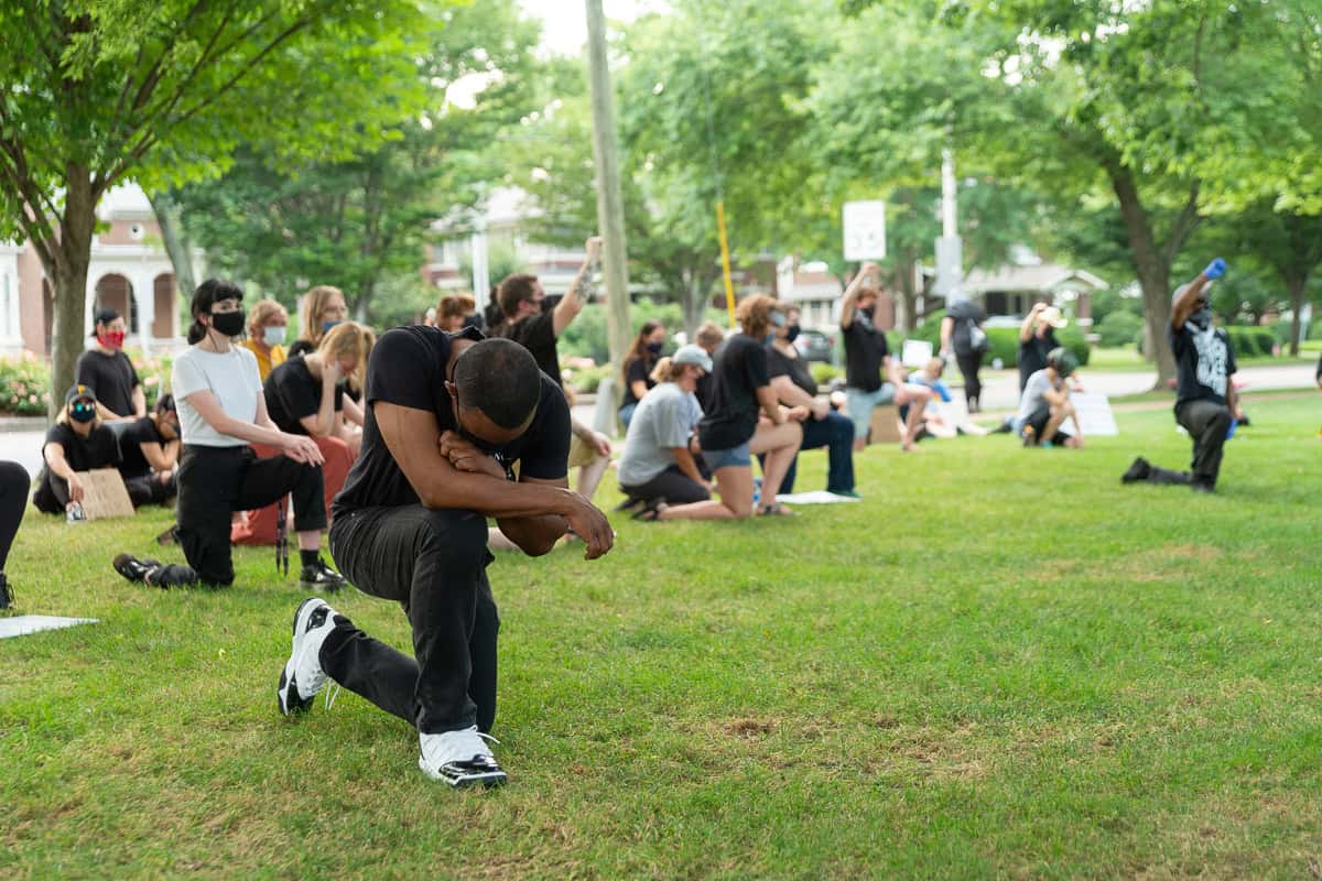 A Black man kneels on one knee in protest with head bowed.