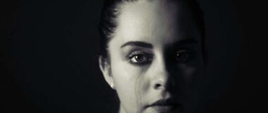 A woman in this black and white image is broad lit and stares ahead blankly with a tear running down her cheek.