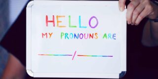 A person holds up a small white board with multi-colored words saying "Hello, my pronouns are..."