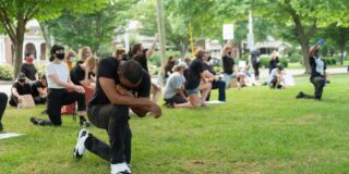 A Black man kneels on one knee in protest with head bowed.