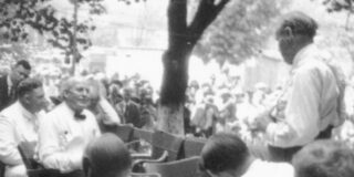 The black and white image shows the outdoor proceedings of the Scopes trial with multiple men dressed in white dress shirts.