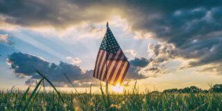 An American flag in a field with the setting sun and clouds in the background.