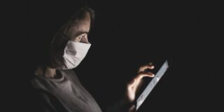 A masked person sits in a very dark room swiping at a phone.
