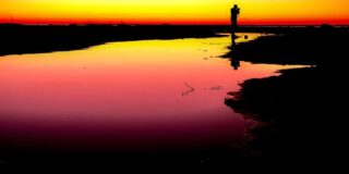 a person stands in silhouette holding a camera against a sunset surrounded by low water as if in a marsh