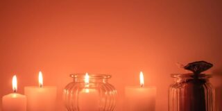 Four short white candles, one in a glass votive, are lit with a soft orange background and whole spices in the foreground.