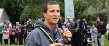 Bear Grylls is shown in casual clothes holding a microphone and speaking at an outdoor event.