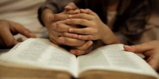 Small hands are clasped and rest on an open bible while adult hands hold the bible's sides.