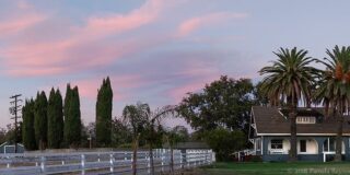 A two story circa 1890's ranch house is shown at the end of a long driveway lined with white fencing at sunset.