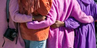 Black women are shown from the back wearing pink and orange with arms around each other's waist.
