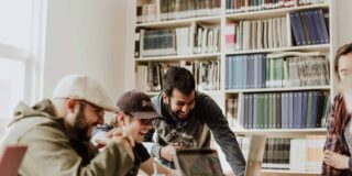 Three men in a library looking at a laptop and laughing.