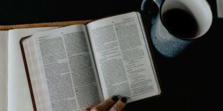 A Bible sits open to Romans with a cup of coffee and a woman's hands visible.