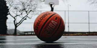 A worn basketball sits on a wet outside court with the hoop behind it.