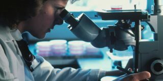 A woman in a white lab coat stares intently into a microscope.